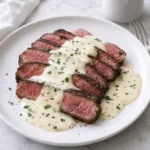 Garlic cream sauce spooned over sliced steak on a white plate with parsley and black pepper